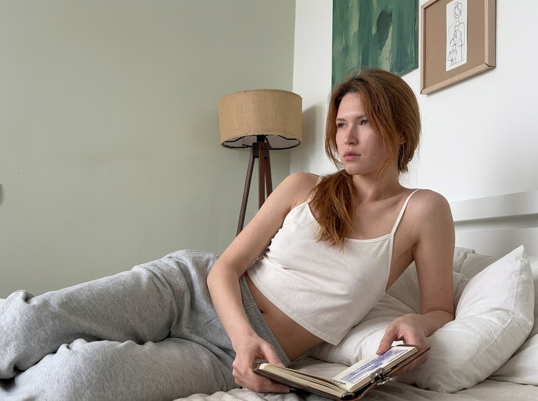 A medium shot of a person wearing a loose, very cropped camisole with thin straps, made of very thin, transparent fabric, with deep armhole cuts. No covering over the garment. The lighting is soft and warm, with a slightly 24-28mm equivalent lens perspective. The overall tone is intimate and casual.