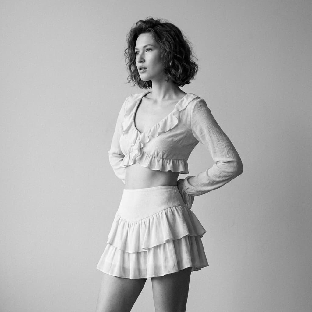 Reference image: conceptual variant. Ultra-realistic black-and-white fashion editorial portrait, studio photography. Vertical format, mid-to-full body framing. A woman standing in three-quarter profile, body slightly turned to the side, one hip subtly shifted back, hands placed behind her lower back. Head turned away from the camera, gaze directed off-frame to the left, calm confident expression, slightly parted lips. Outfit: delicate two-piece fashion set in light fabric. Cropped long-sleeve top with ruffled edges and a deep front opening, paired with high-waisted layered ruffle mini skirt. Outfit is styled as high-fashion editorial wear, elegant and intentional, not casual, not lingerie-focused. Styling: short wavy bob haircut, softly tousled, natural volume. Makeup minimal and editorial: smooth skin, subtle contour, defined eyes, glossy lips. No accessories, no jewelry, no additional styling elements. Lighting: soft studio light, evenly diffused, gentle shadows sculpting the body and face. High contrast black-and-white tonality with fine film grain texture. No harsh highlights, no blown whites. Background: plain neutral studio backdrop, seamless, clean, no texture, no props. Mood: elegant, confident, timeless, classic fashion editorial. Calm body language, controlled posture, refined attitude. Camera: professional studio camera, 85mm lens look, shallow depth of field, sharp focus on face and torso. Style: Vogue-style editorial, minimalist fashion photography, cinematic realism. Strict rules: black and white only, no color tones, no pose change, no outfit reinterpretation, no added accessories, no environment changes, no exaggerated anatomy, no sexual framing, fashion editorial context only.
